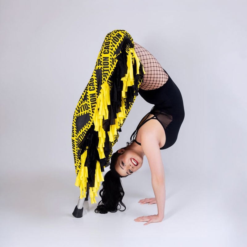 Woman in black leotard, fishnet stockings, handstand backbend, yellow caution tape fabric, smiling.