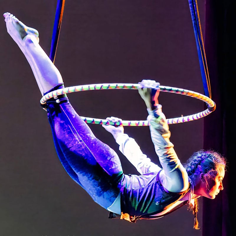 Female aerialist in blue outfit performs on a colorful aerial hoop, legs extended upward, suspended by straps.