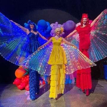 Three stilt walkers in vibrant dresses with illuminated wings and balloons on stage