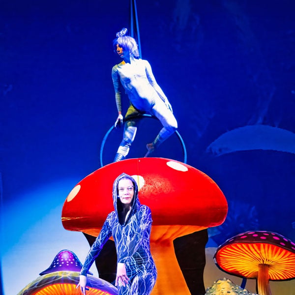 Two costumed performers on a stage with giant red and glowing mushrooms, one balancing on a hoop atop a red mushroom.