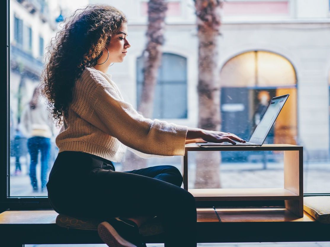 A woman with curly hair sits by a window, using a laptop on a small wooden table.
